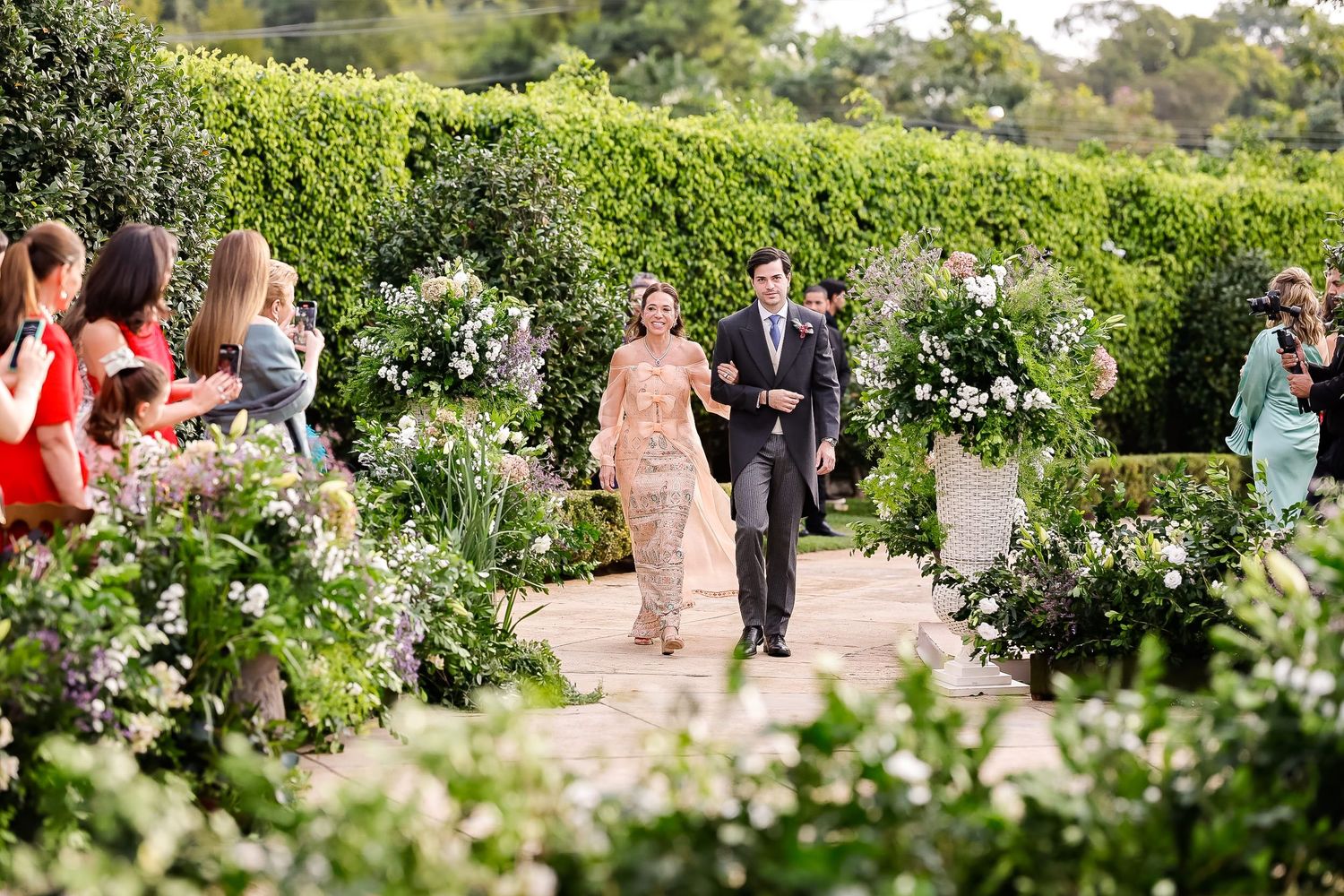 Wedding guests walk through a lush garden path lined with blooming white flowers and greenery on a sunny day.