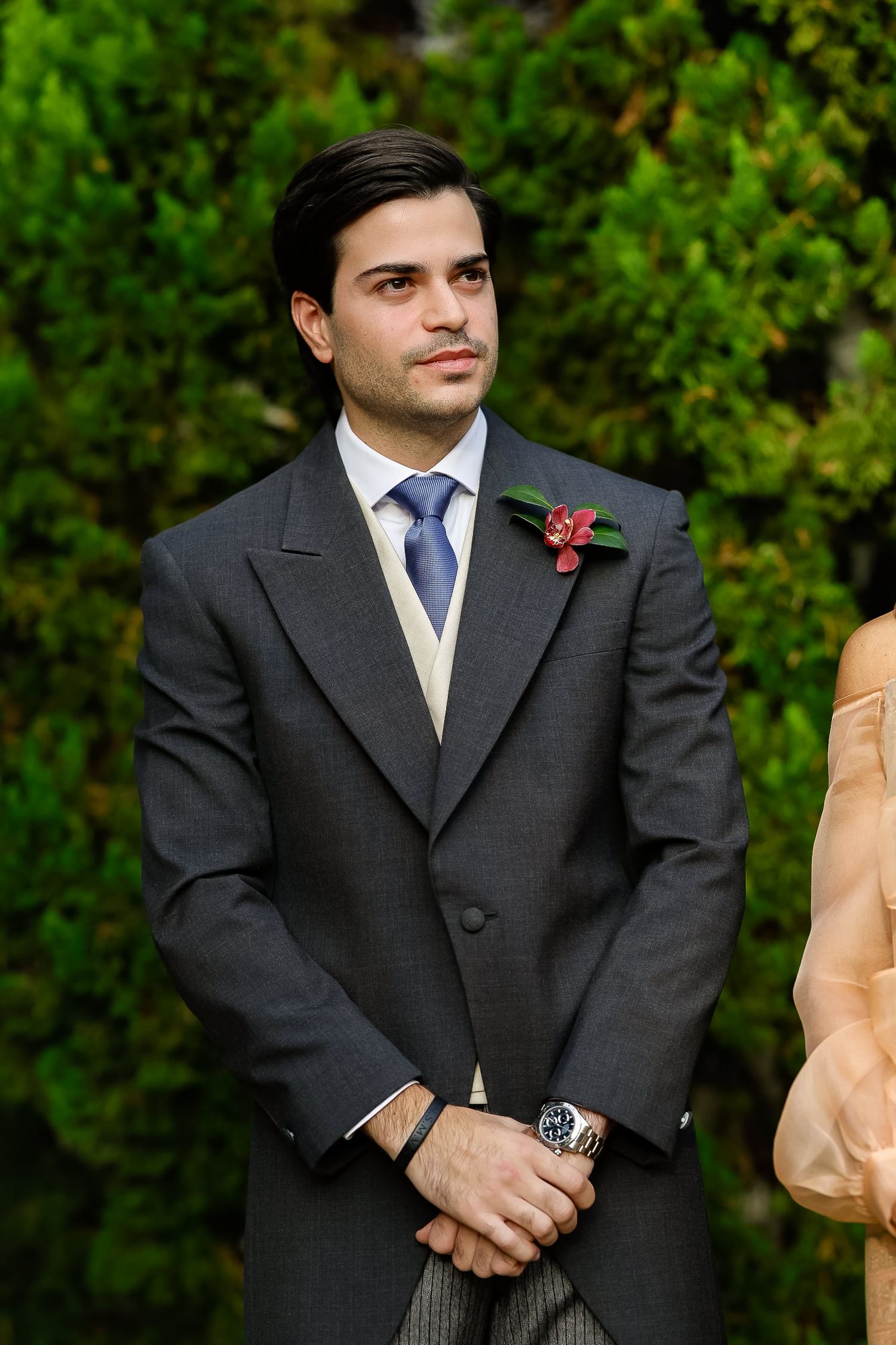A groomsman in a dark suit with a red boutonniere stands during an outdoor wedding ceremony.