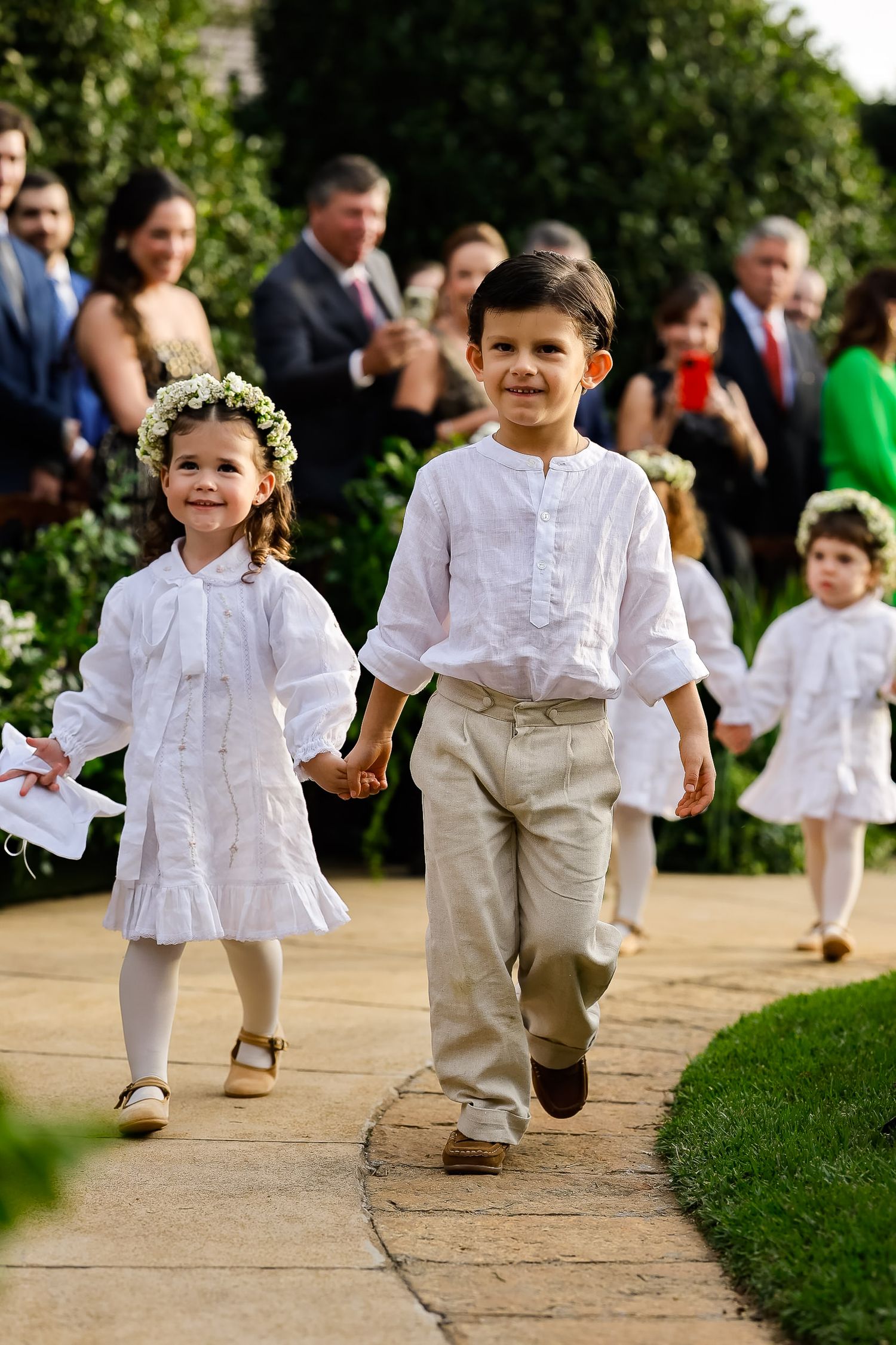 Children in white shirts and khaki pants run playfully along a garden path during a formal outdoor event.