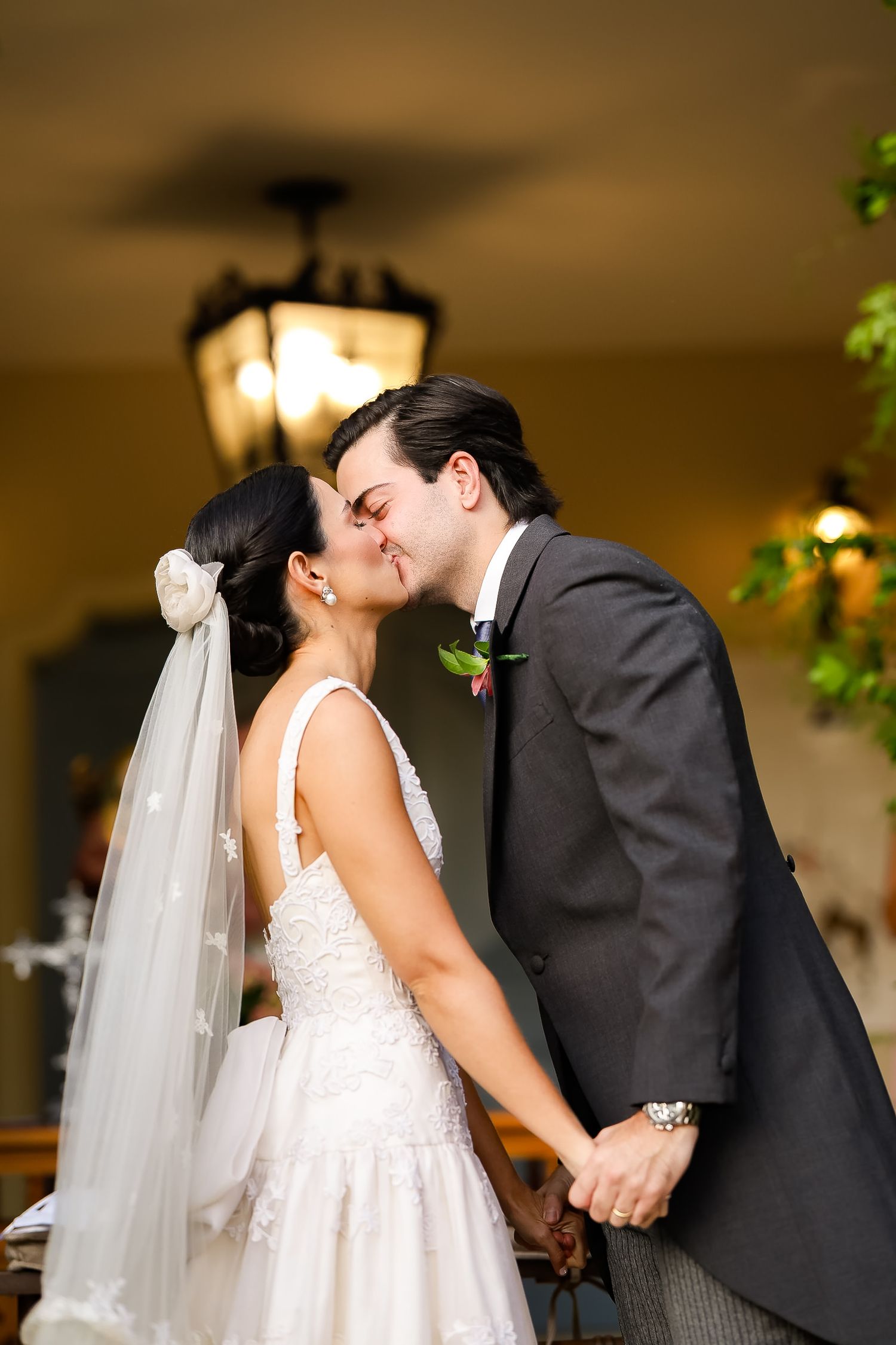 A bride in a white lace gown and veil shares a romantic kiss with the groom during their wedding celebration.