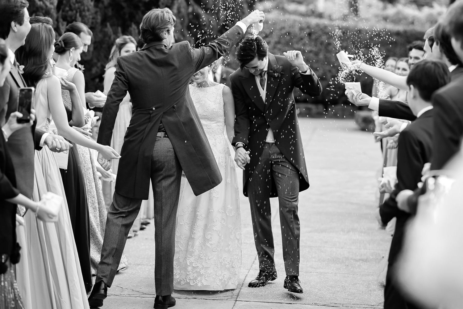 A black and white wedding celebration photo showing guests joyfully tossing confetti at a couple during their outdoor ceremony.