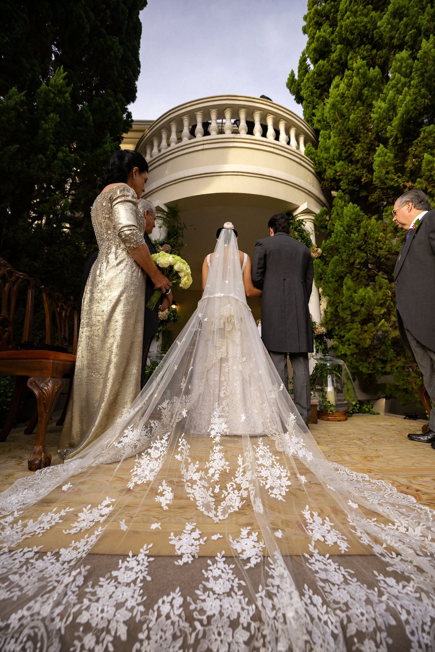 Stunning cathedral-length wedding veil with intricate lace details trails behind bride and groom in outdoor garden setting.