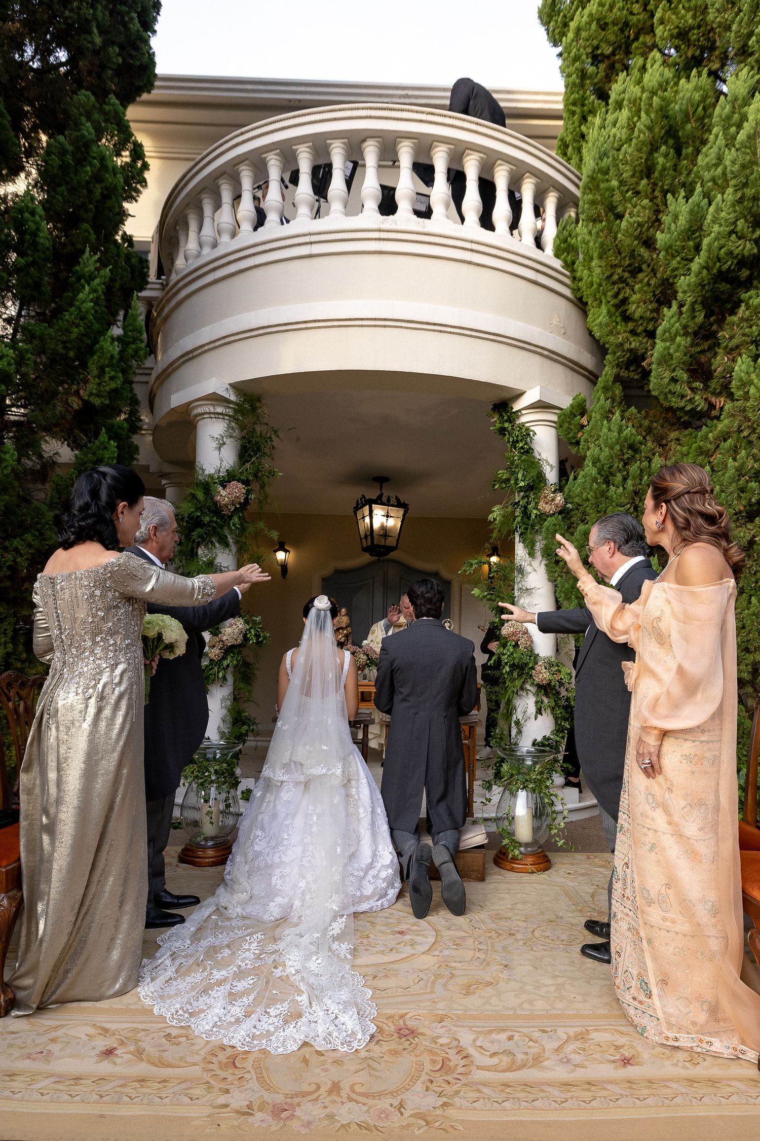 Wedding party stands under an elegant arched entrance with white columns and lush greenery during outdoor ceremony.