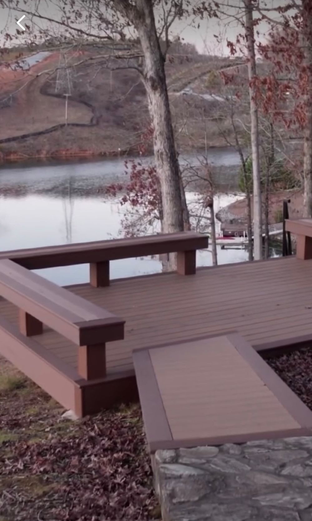 A wooden deck with built-in benches overlooks a serene lake surrounded by autumn trees and fallen leaves.