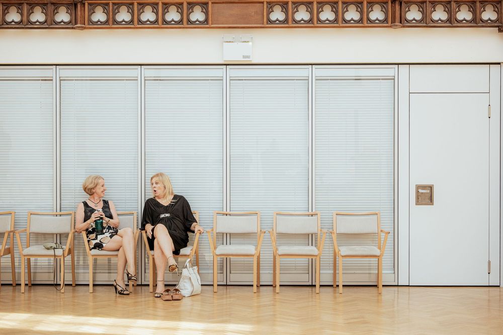 Two people sit together on wooden chairs against a light blue wall in a minimalist interior setting.