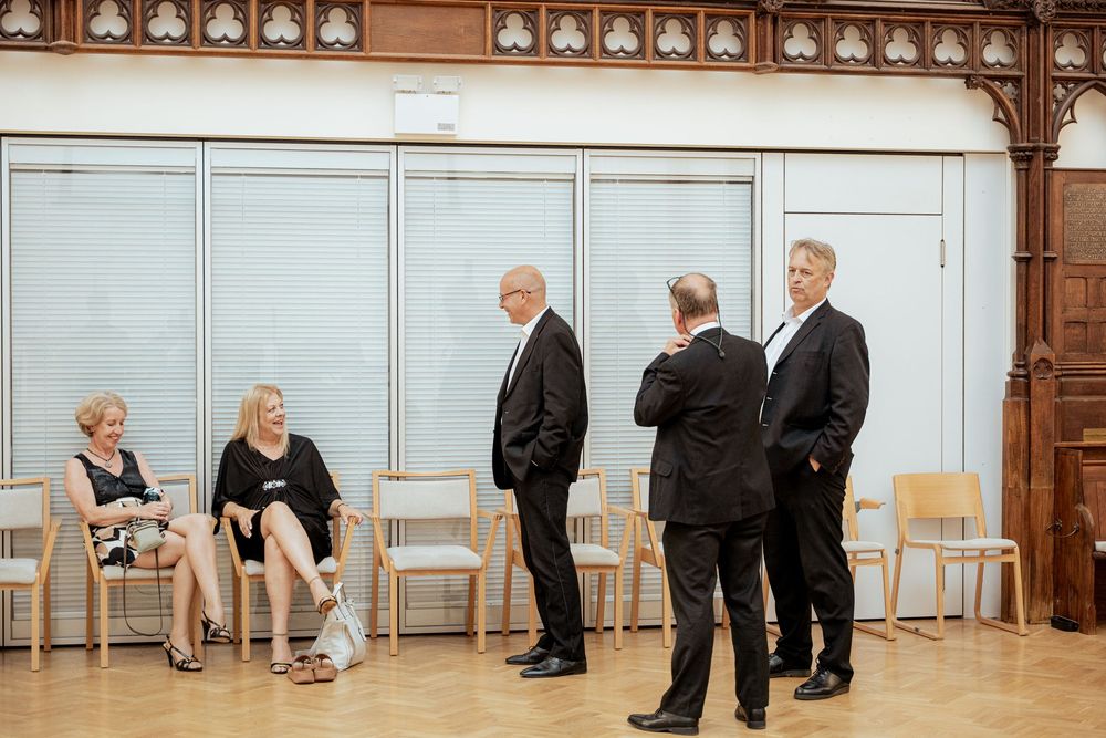 People stand and sit along a wall in a formal event space with wooden paneling and white walls.