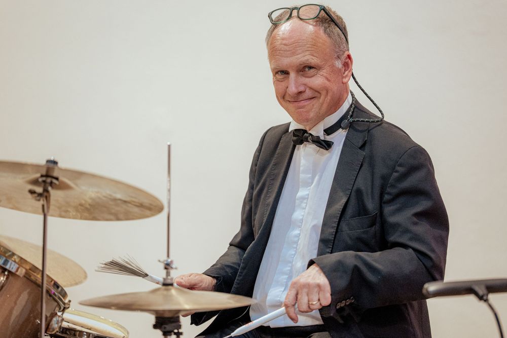 Drummer in formal attire plays on a drum kit with cymbals during a musical performance.