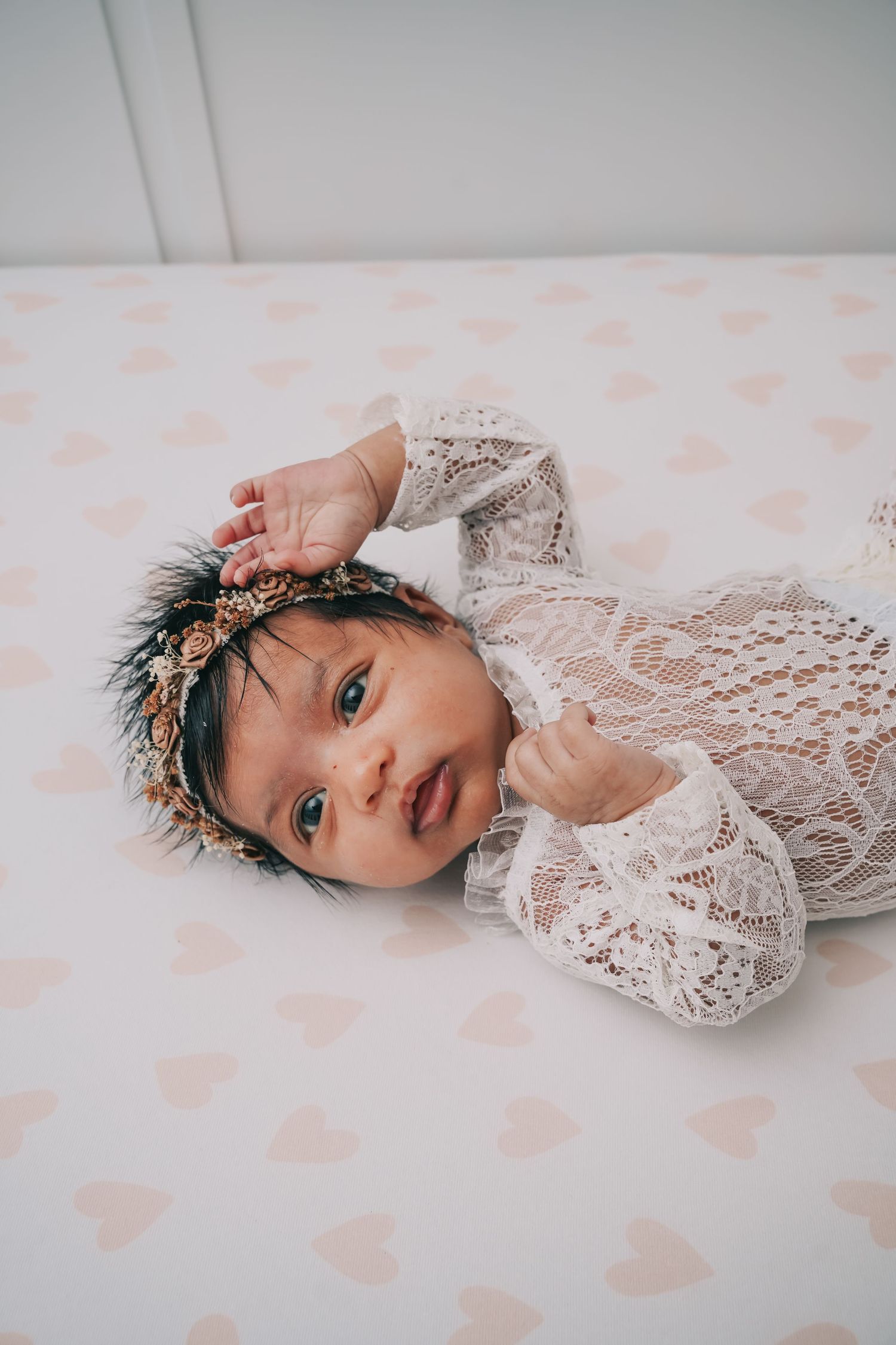 Newborn baby wearing delicate white lace dress and floral crown lying on soft white blanket with pink heart pattern.