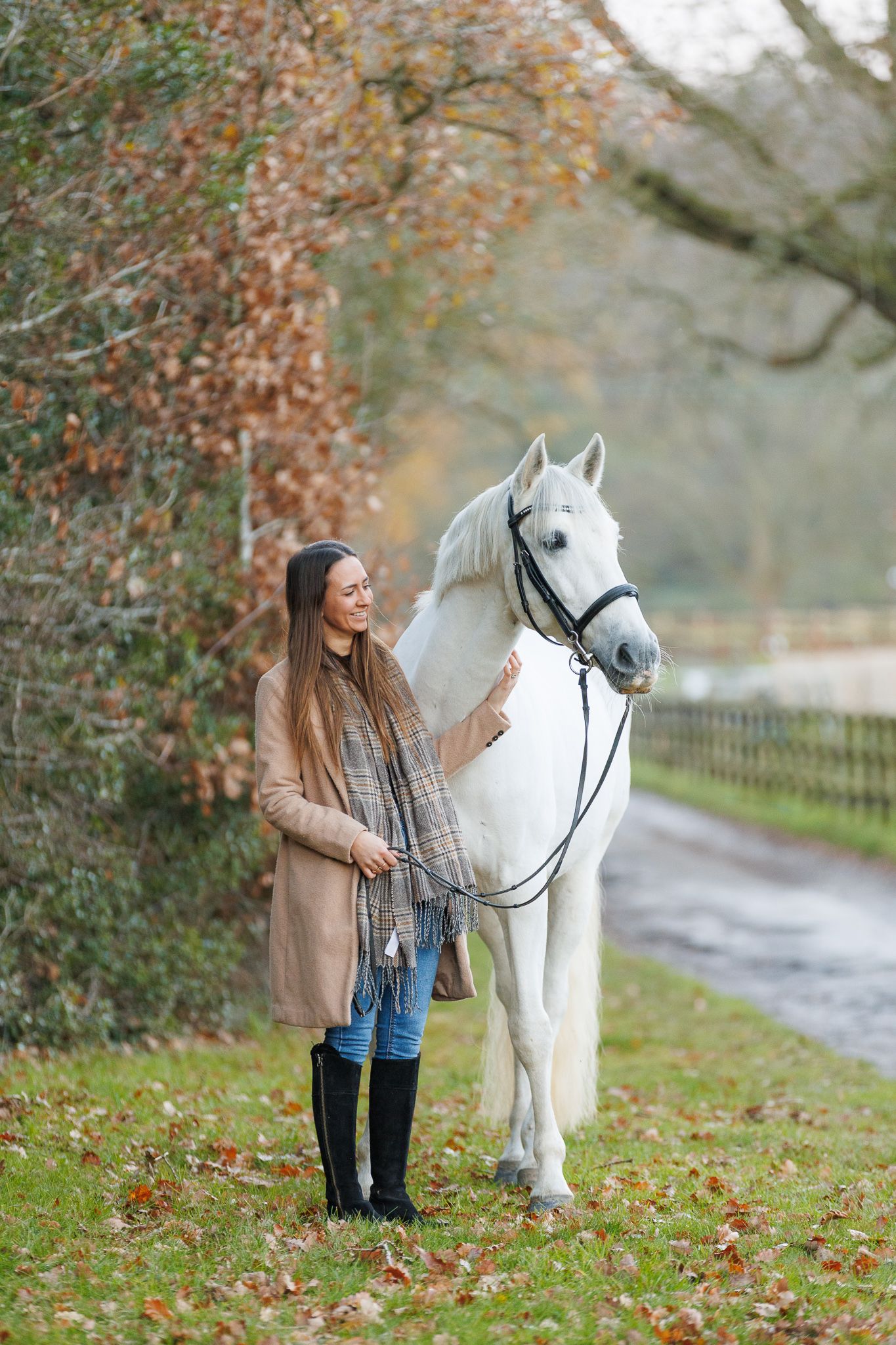 A beautiful white horse stands with its owner on an autumn path surrounded by fallen leaves and trees.