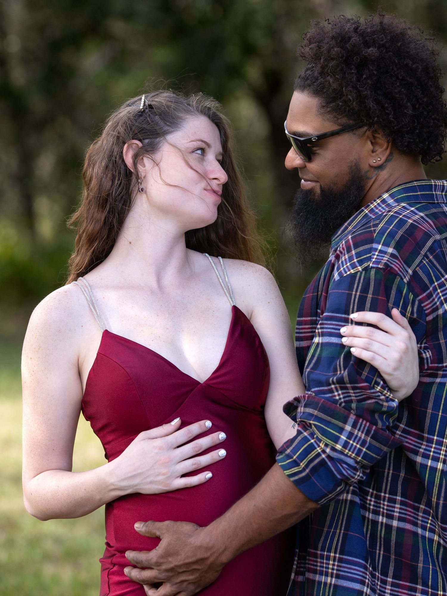A couple in a burgundy dress and plaid shirt embrace lovingly on a grassy field under trees.