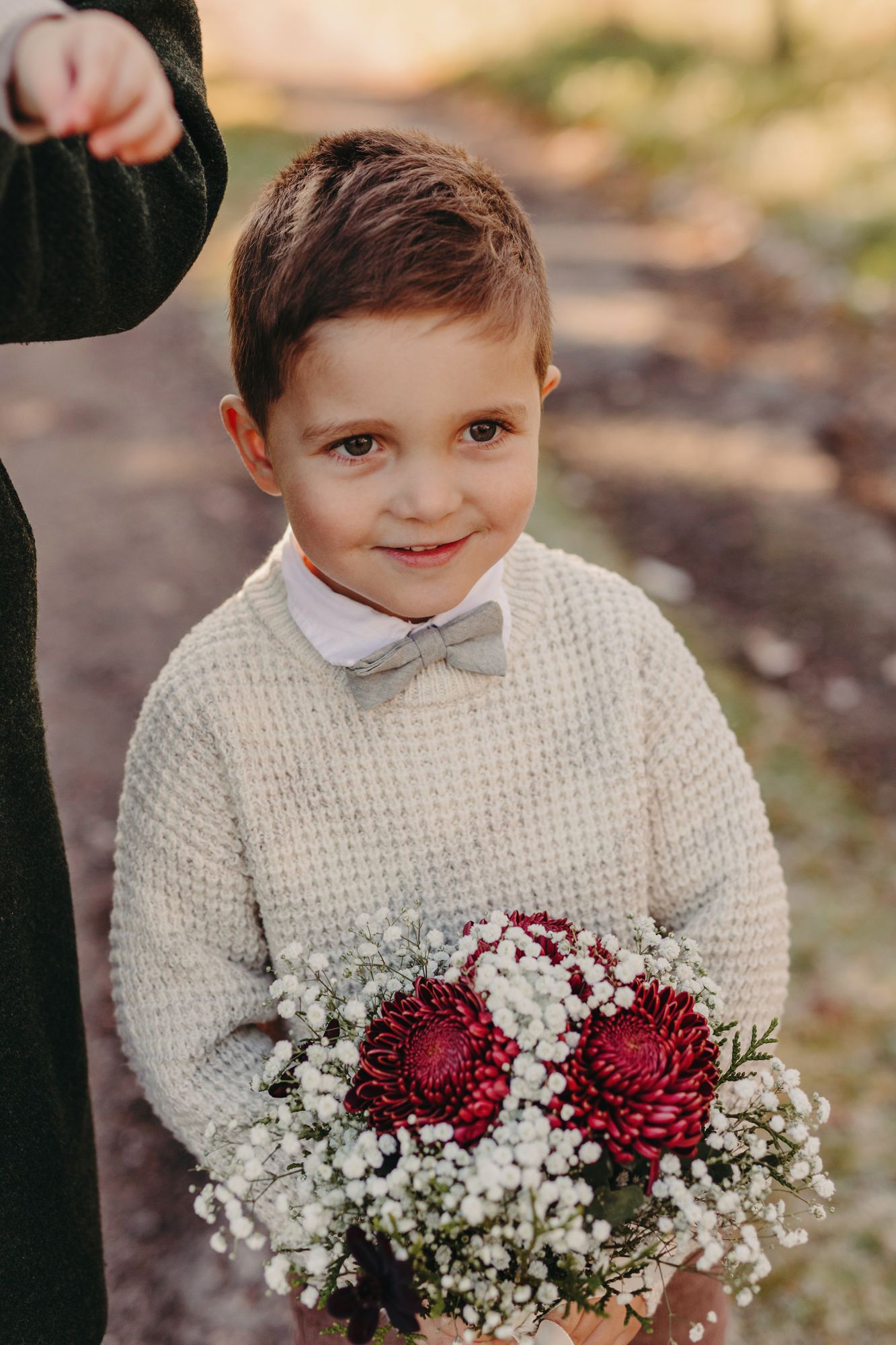 A young child in a cream knit sweater holds a beautiful bouquet of burgundy dahlias and baby's breath flowers.