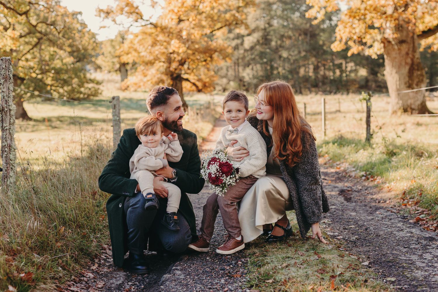 A couple sits together on a path surrounded by autumn trees with golden leaves during a fall photo session.