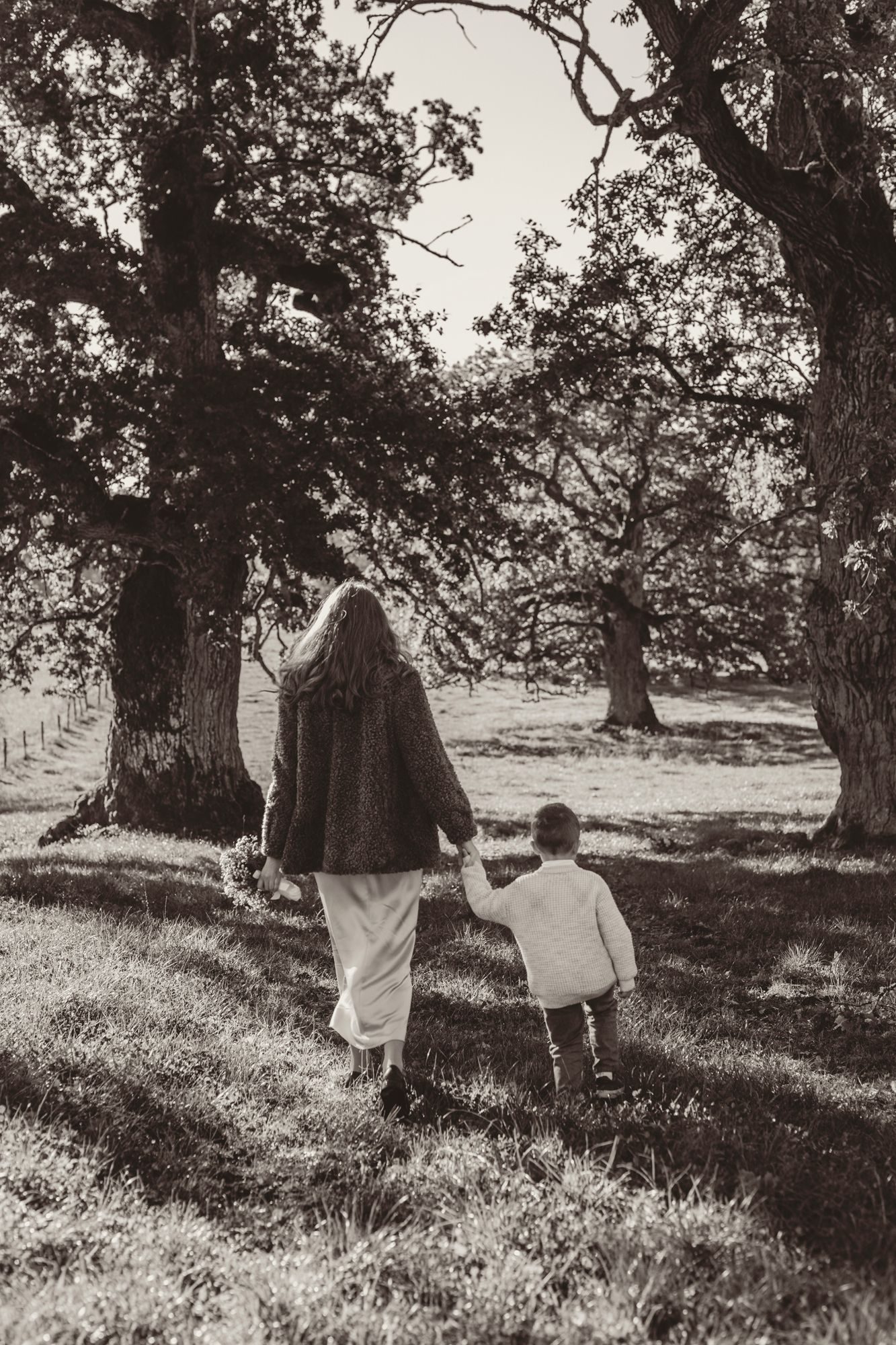 Black and white photo of two people walking together through a sunlit grove of trees on a grassy hillside.