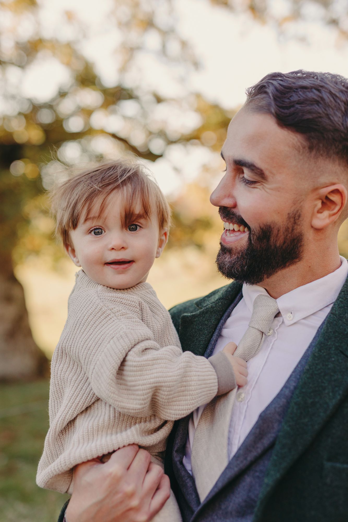 A father in a dark blazer and tie smiles while holding a toddler wearing a beige sweater during golden hour outdoors.