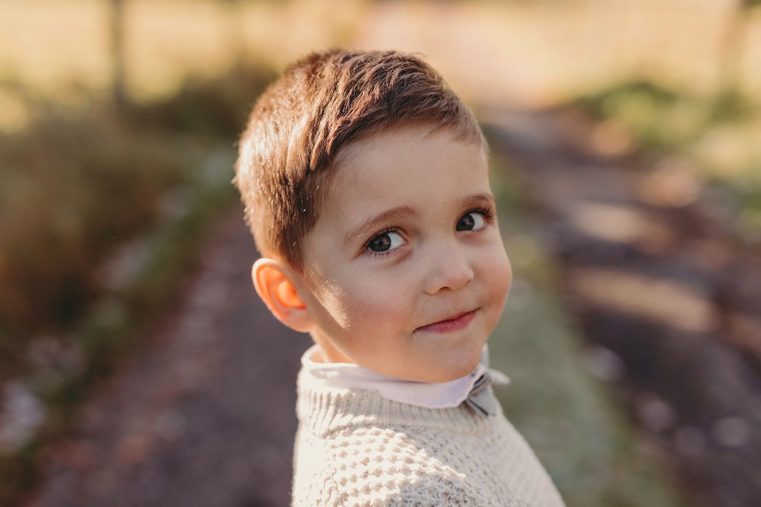 A young child wearing a white knit sweater gazes upward with a thoughtful expression in an outdoor setting.