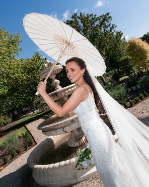 Bride in white gown holding parasol umbrella beside stone garden fountain on sunny day.