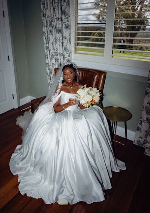 A person in a flowing white wedding gown sits elegantly on a brown leather chair in a sunlit room with floral curtains.