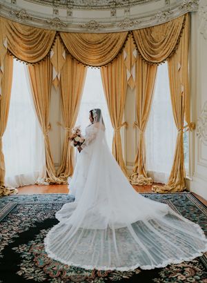 Bride in a white wedding gown with long veil stands before golden draped windows in an ornate ballroom.