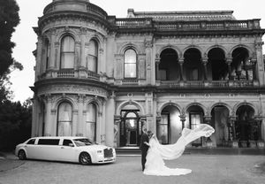 Bride and groom embrace before a grand Victorian mansion with a white stretch limousine in a black and white wedding photo.