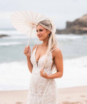 A person in a white lace wedding dress holds a light pink parasol while standing on a beach with ocean waves behind.