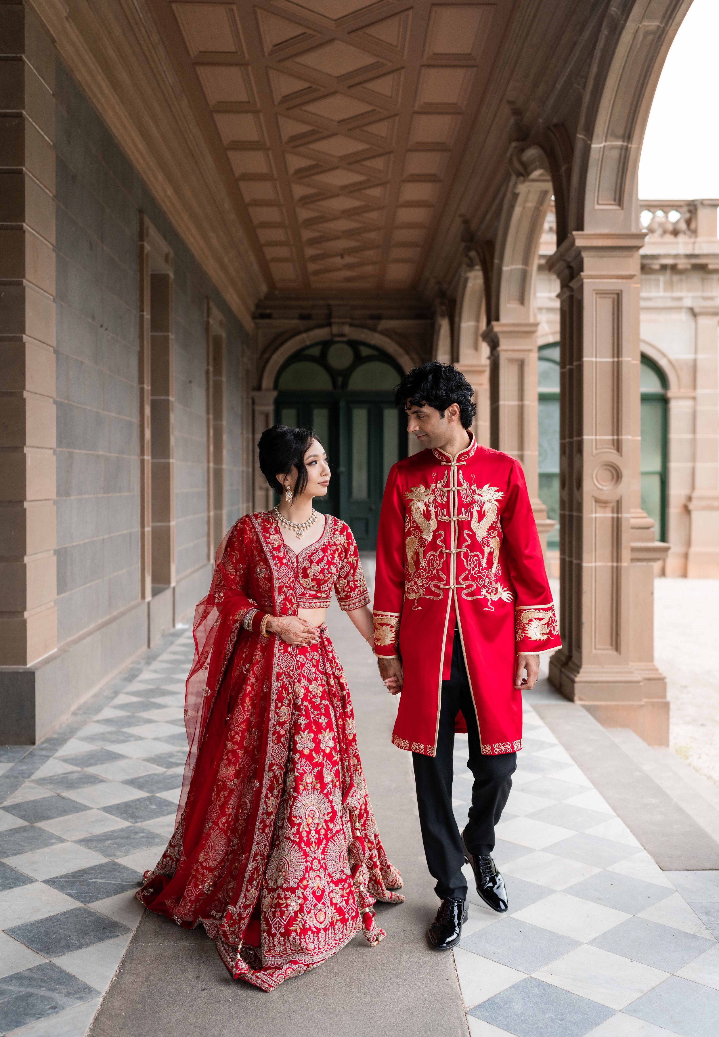 Couple in traditional red wedding attire walking together in an elegant stone hallway with arches and checkered floor.
