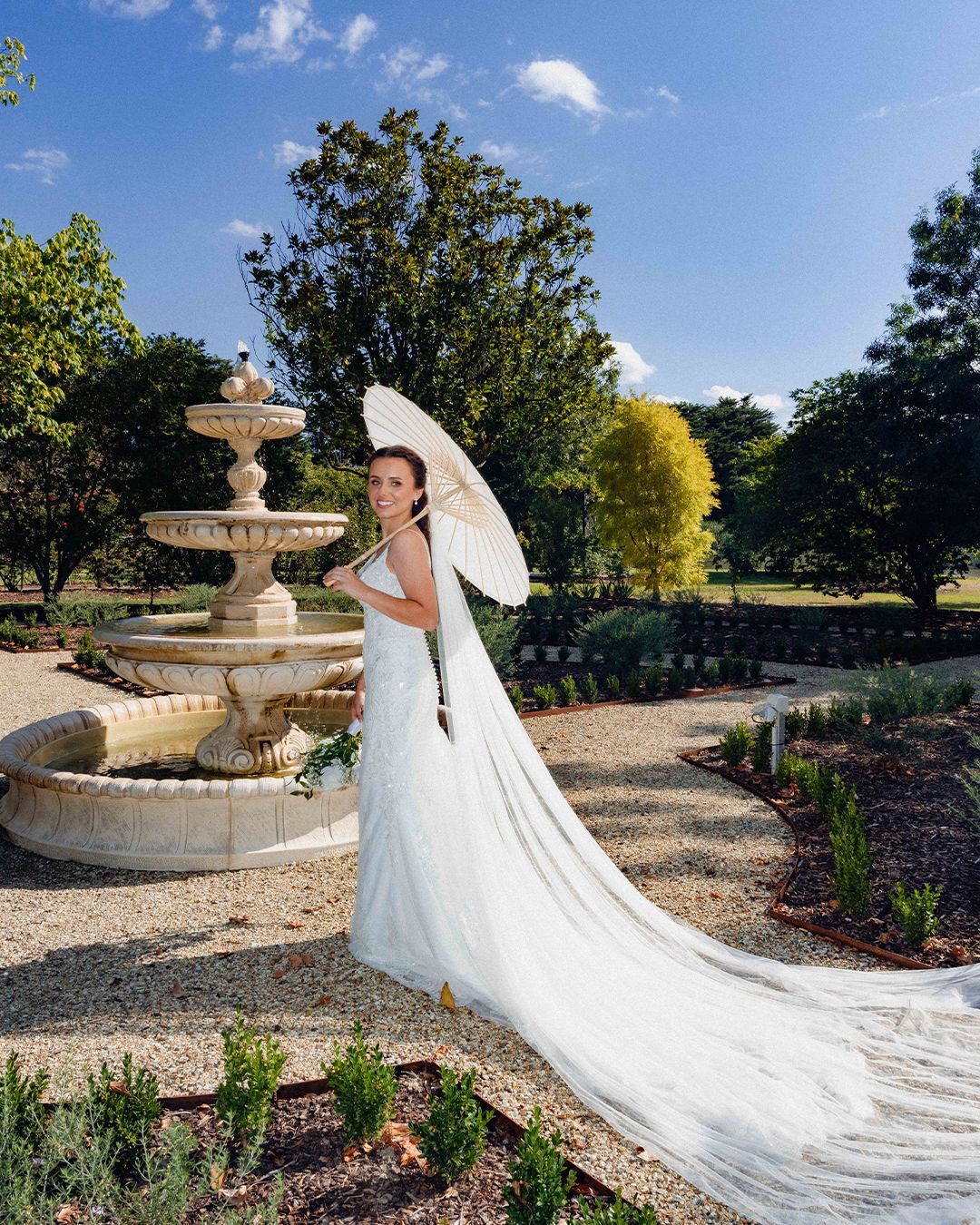 Bride in white gown holding parasol stands by stone fountain in lush garden on sunny day.