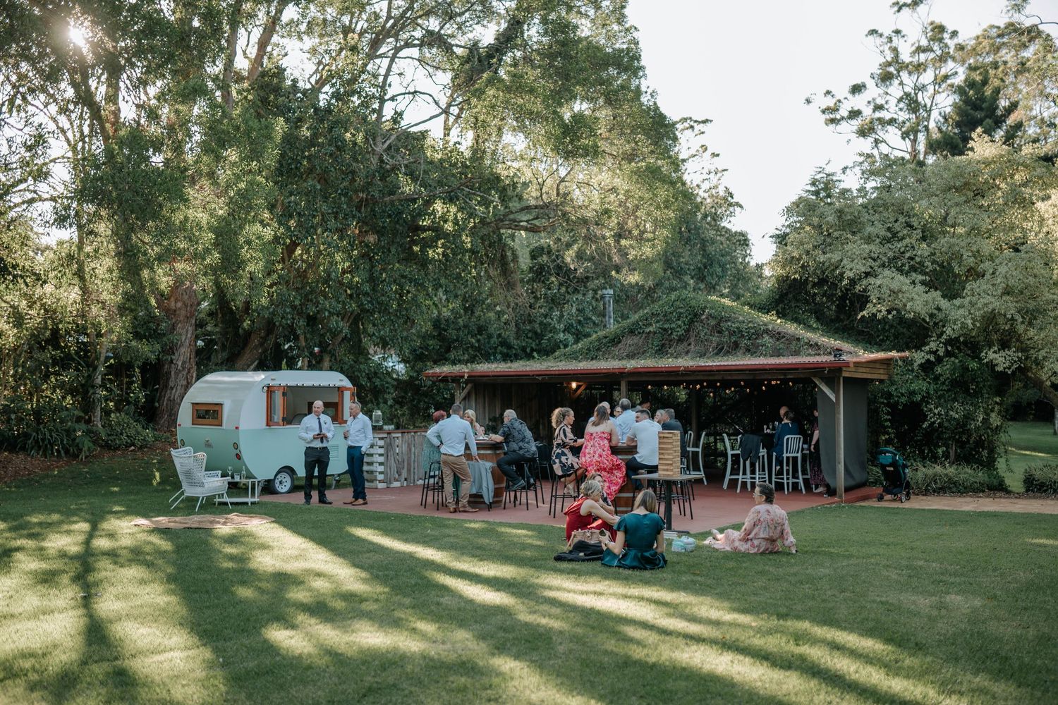 Scenic outdoor wedding venue with golden sunlight streaming through trees onto a grassy lawn with scattered seating.