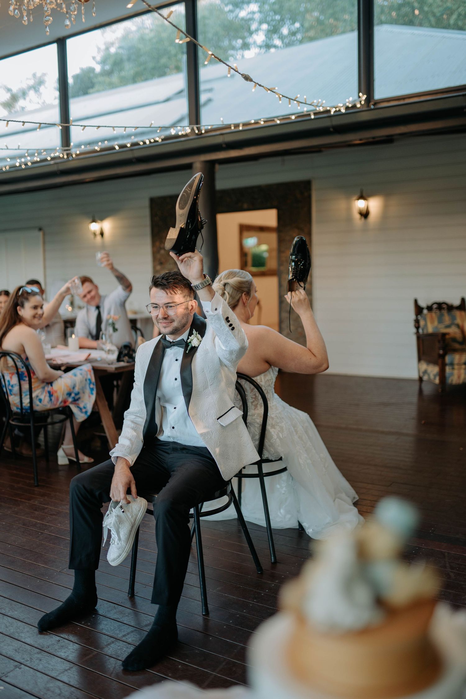 Couple plays wedding shoe game at rustic indoor reception venue with string lights and wooden floors.