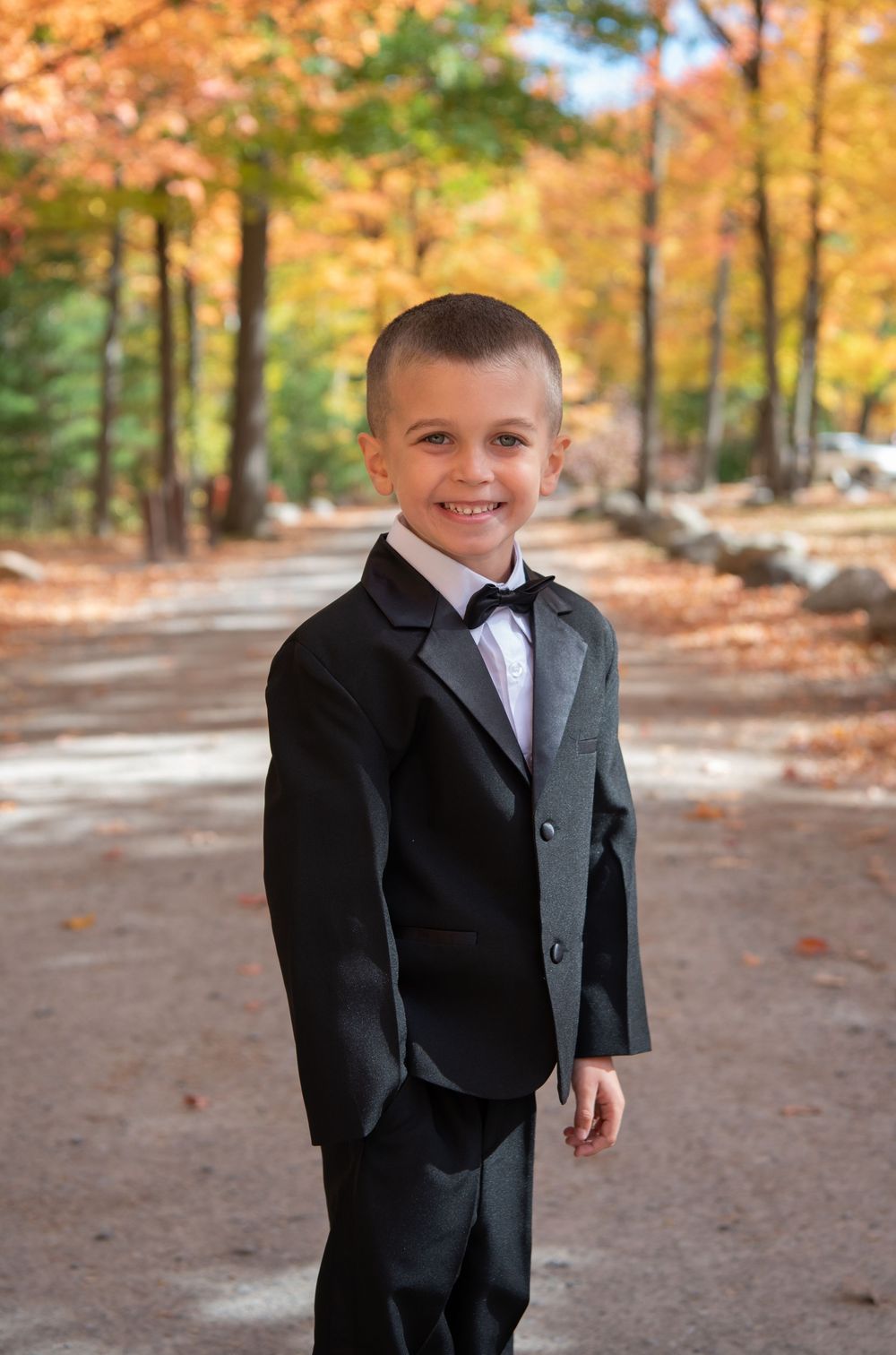 A child wearing a formal black tuxedo stands smiling on a path surrounded by vibrant autumn foliage.