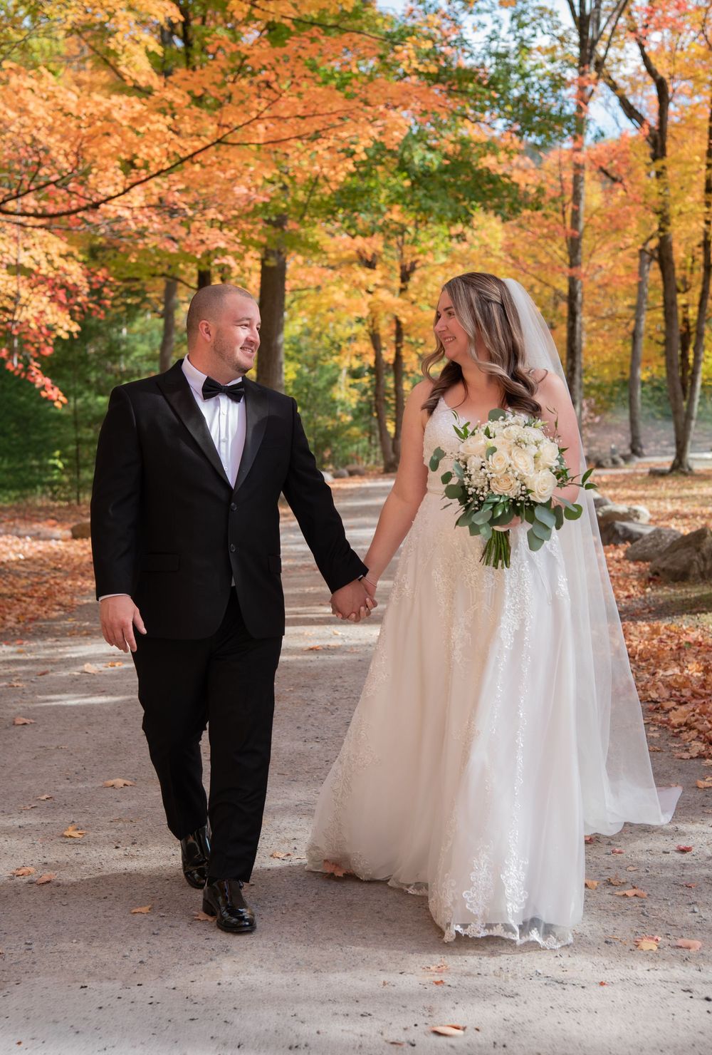 A couple in wedding attire shares a romantic moment on a path surrounded by vibrant autumn foliage.