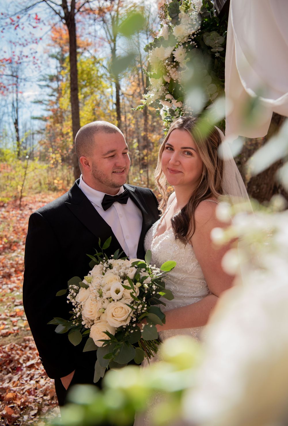 A couple poses for their wedding photos in a fall forest setting with white floral bouquet and formal attire.