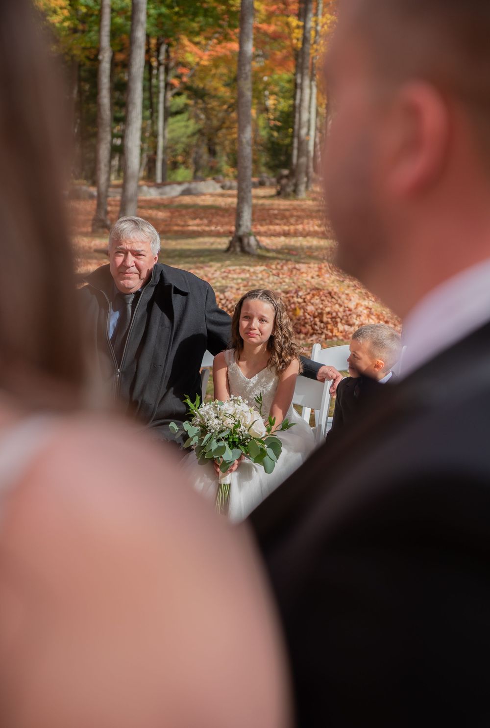 Wedding ceremony guests seated outdoors among autumn trees with fallen leaves on the ground.