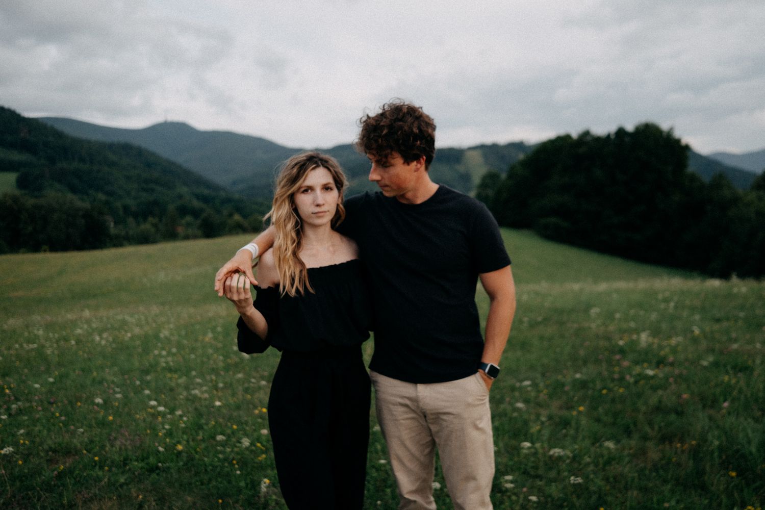 Couple in casual attire standing together in a mountain meadow.