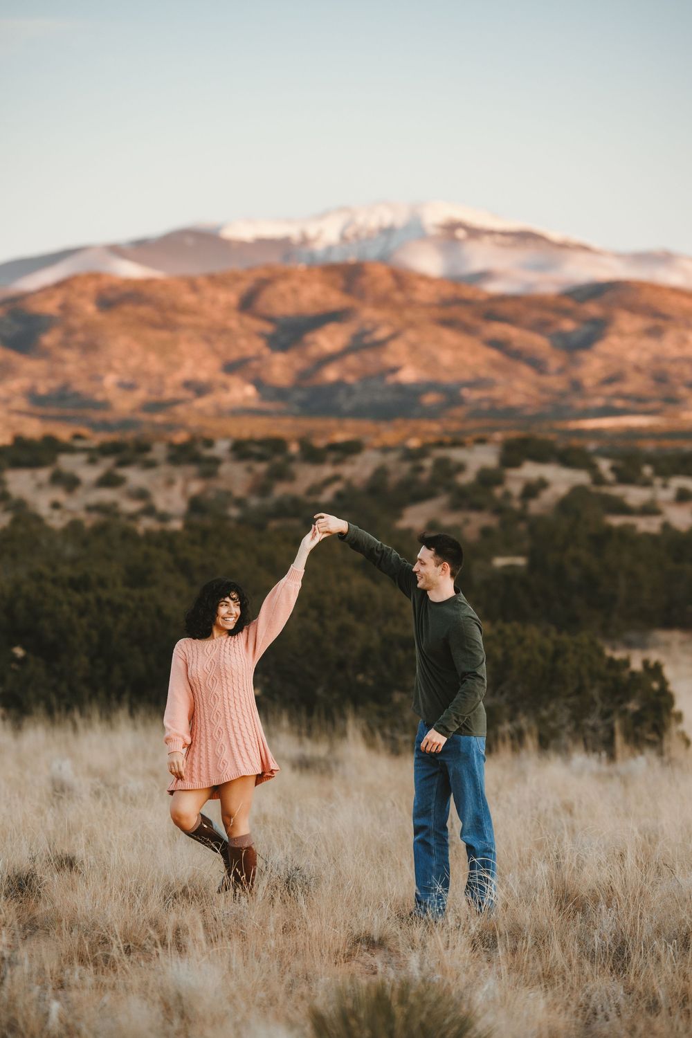 Joyful couple dancing in open field with stunning snow-capped mountain range at sunset.