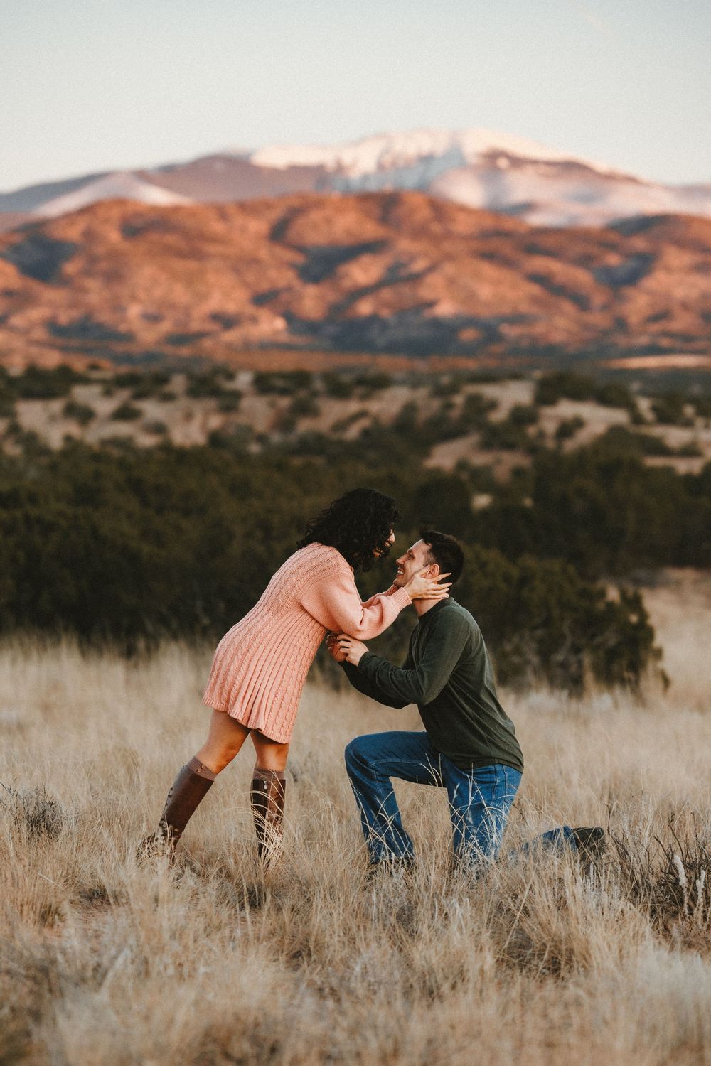 Emotional marriage proposal moment with man on one knee kissing woman in scenic mountain field.