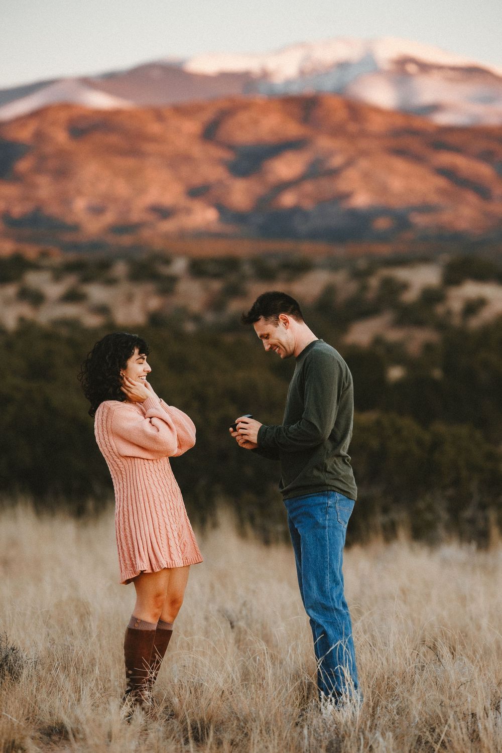 Couple admiring engagement ring together while standing in golden field with mountain backdrop.