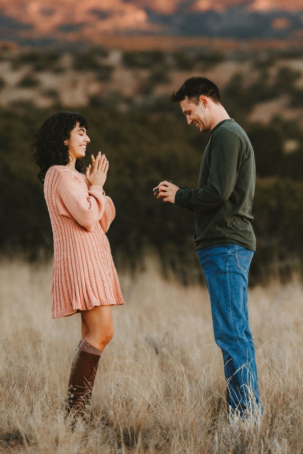 Man presenting engagement ring to surprised woman with clasped hands in mountain landscape.