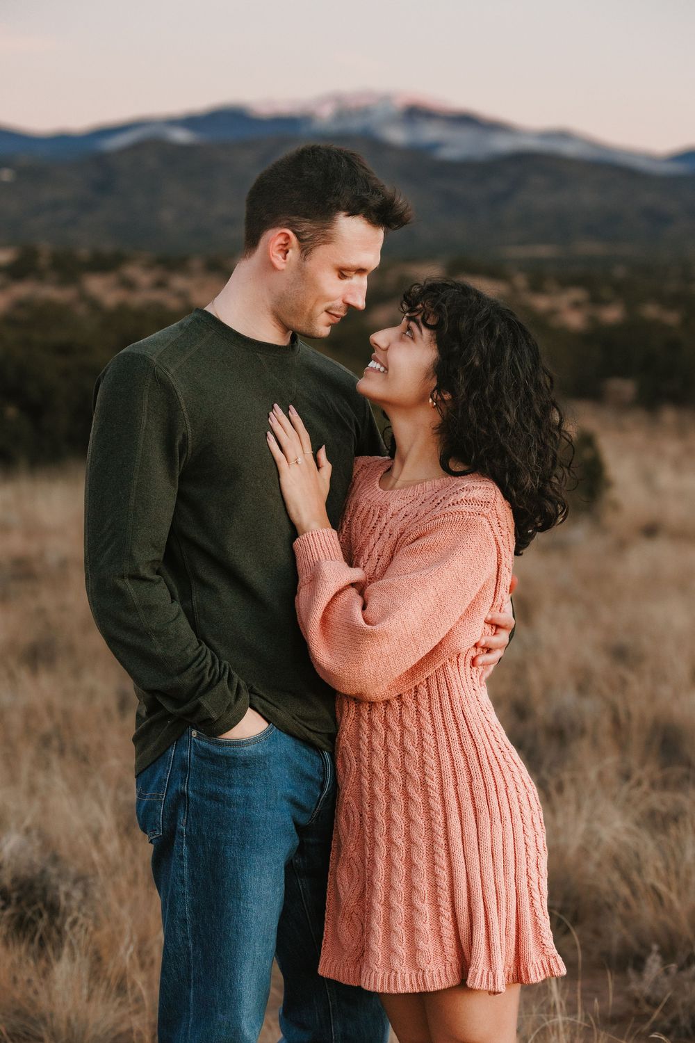 Couple embracing in a field with snow-capped mountains at sunset.