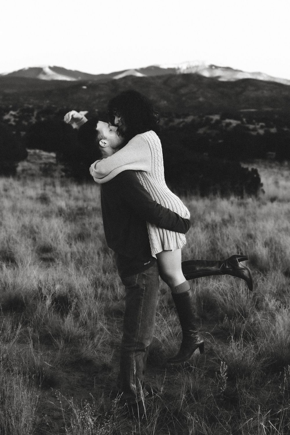 Black and white photo of man lifting woman in a grassy mountain field.