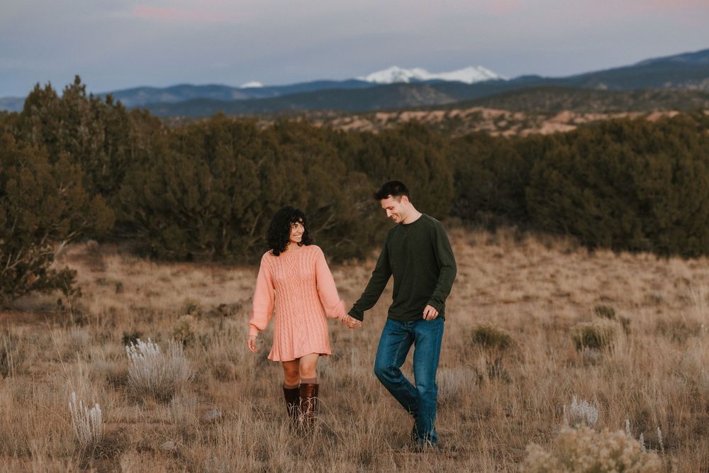 Couple walking hand-in-hand through a dry field with mountains at sunset.
