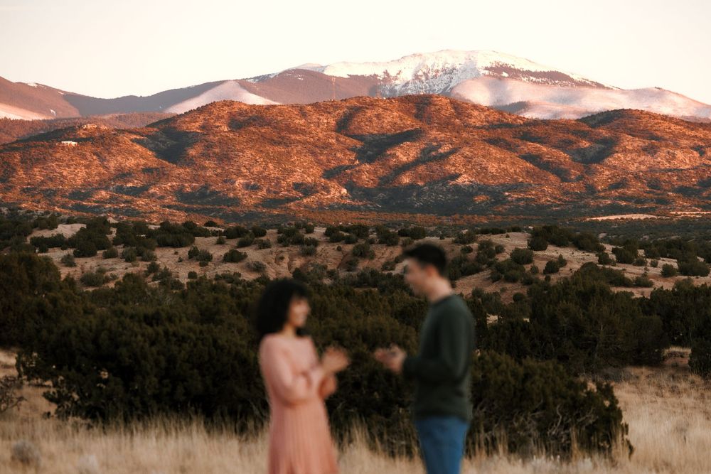 Couple standing in field with stunning snow-dusted mountain backdrop at dusk.