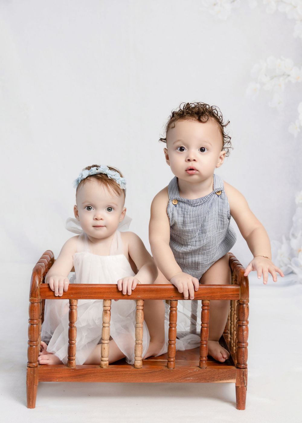 Two babies in a wooden toy crib, girl in white dress with bow headband, boy in gray romper, white studio background.