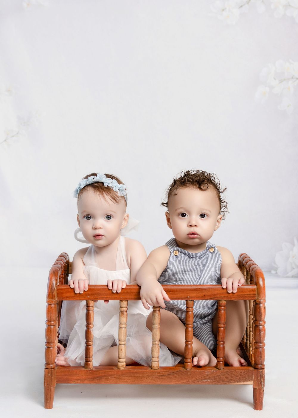 Two adorable babies sit together in a wooden doll crib during a professional photo session.