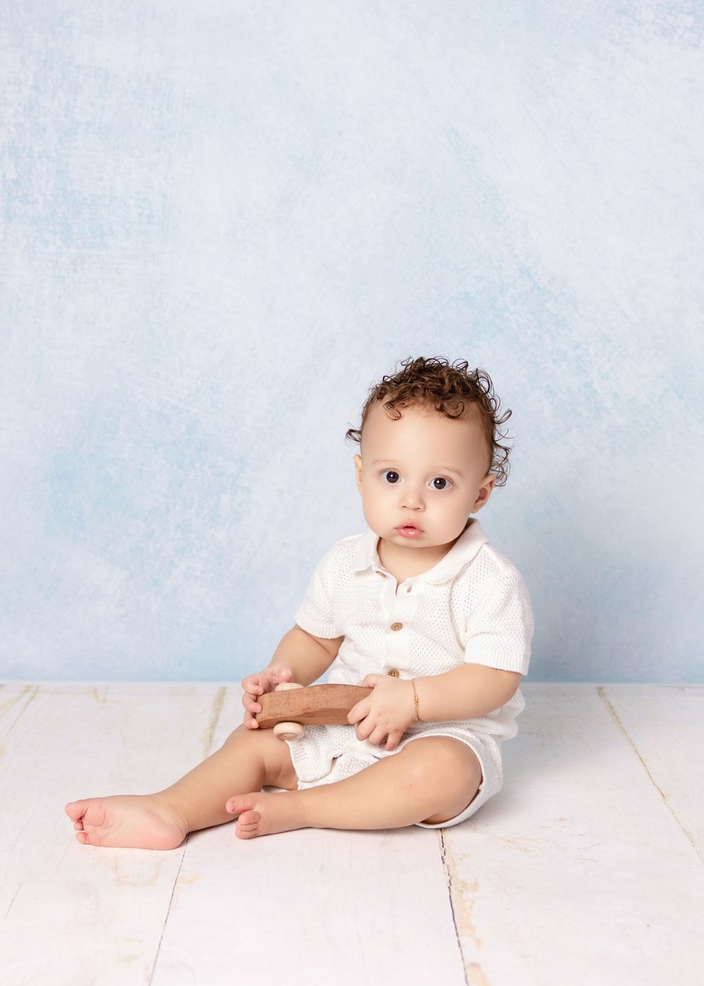 Cute curly-haired baby sits on white floor holding a toy in a white romper during photoshoot.