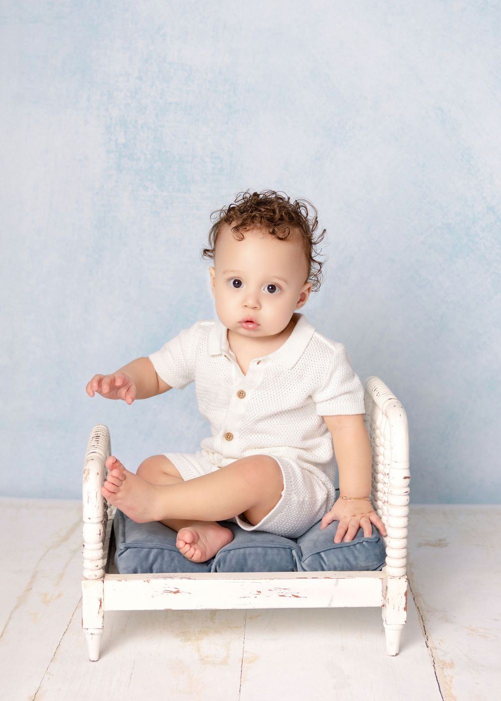 Curly-haired baby in white romper sitting on a miniature white wooden bed against a soft blue backdrop.