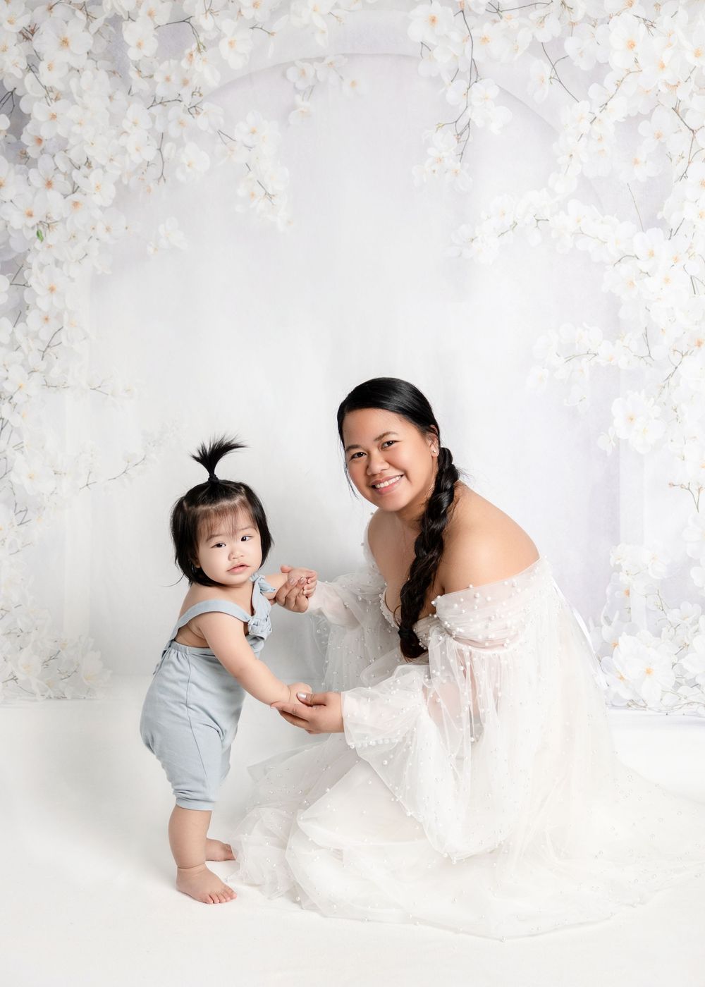 A smiling mother in a white lace gown holds hands with her baby girl in a floral studio setting.