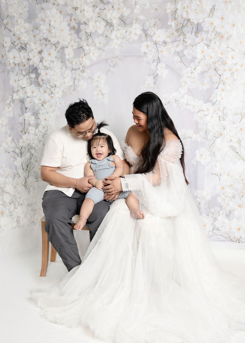 A happy family of three poses together in a white floral studio, parents smiling at their laughing baby.