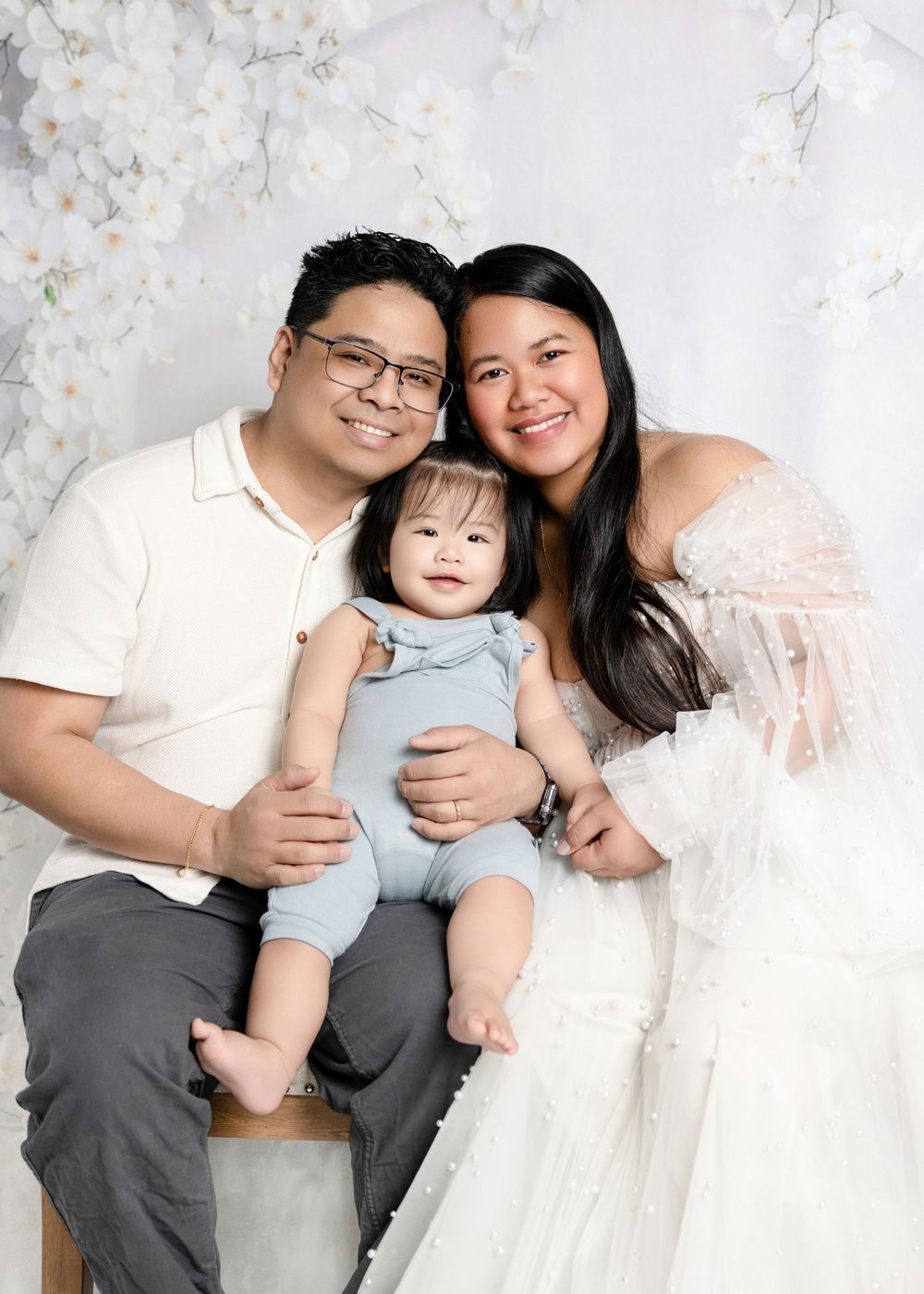 A happy Asian family with a smiling baby in a blue romper poses for a professional studio portrait with floral backdrop.