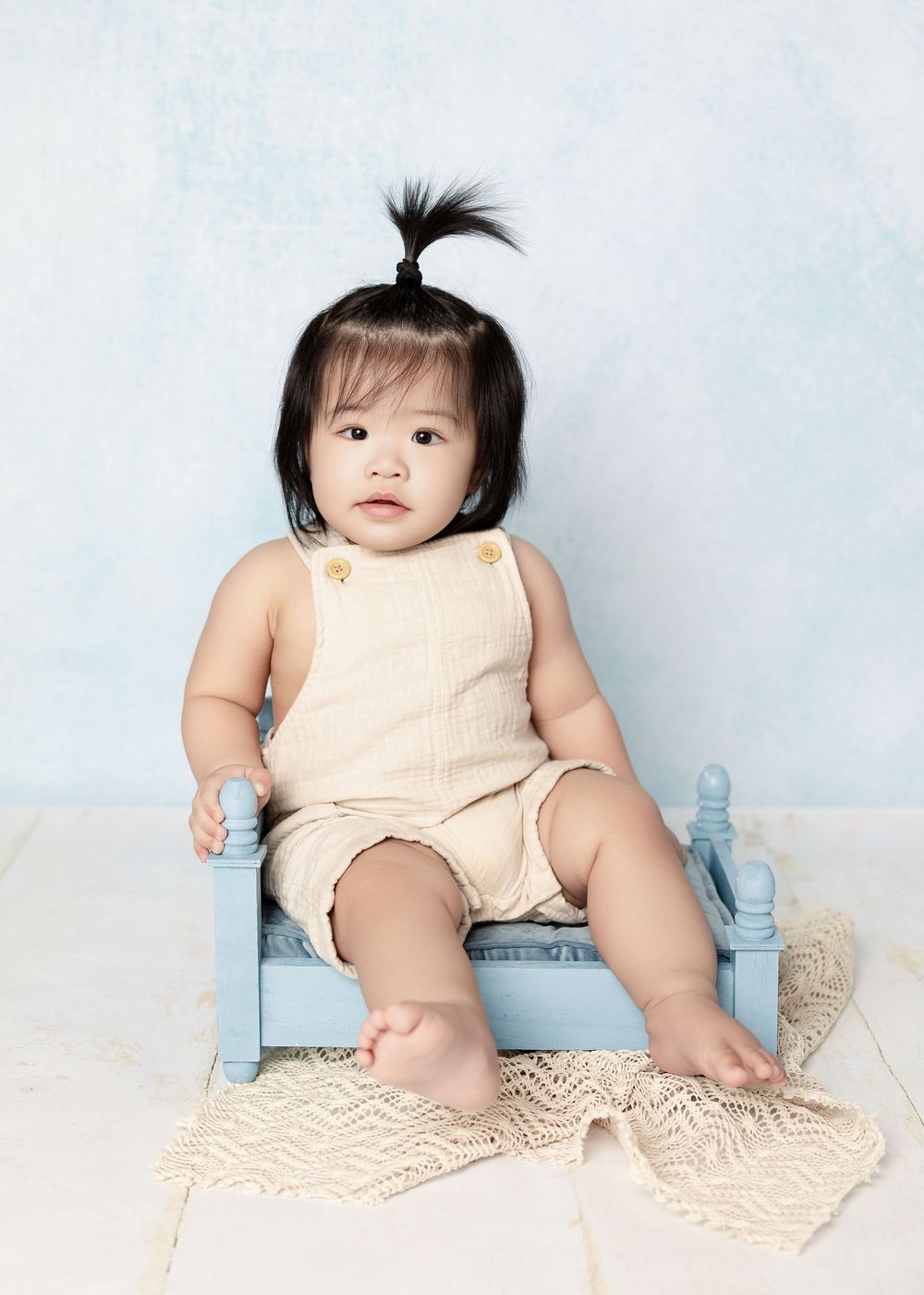 Adorable Asian baby girl with a tiny ponytail sits on a small blue chair wearing a beige romper.