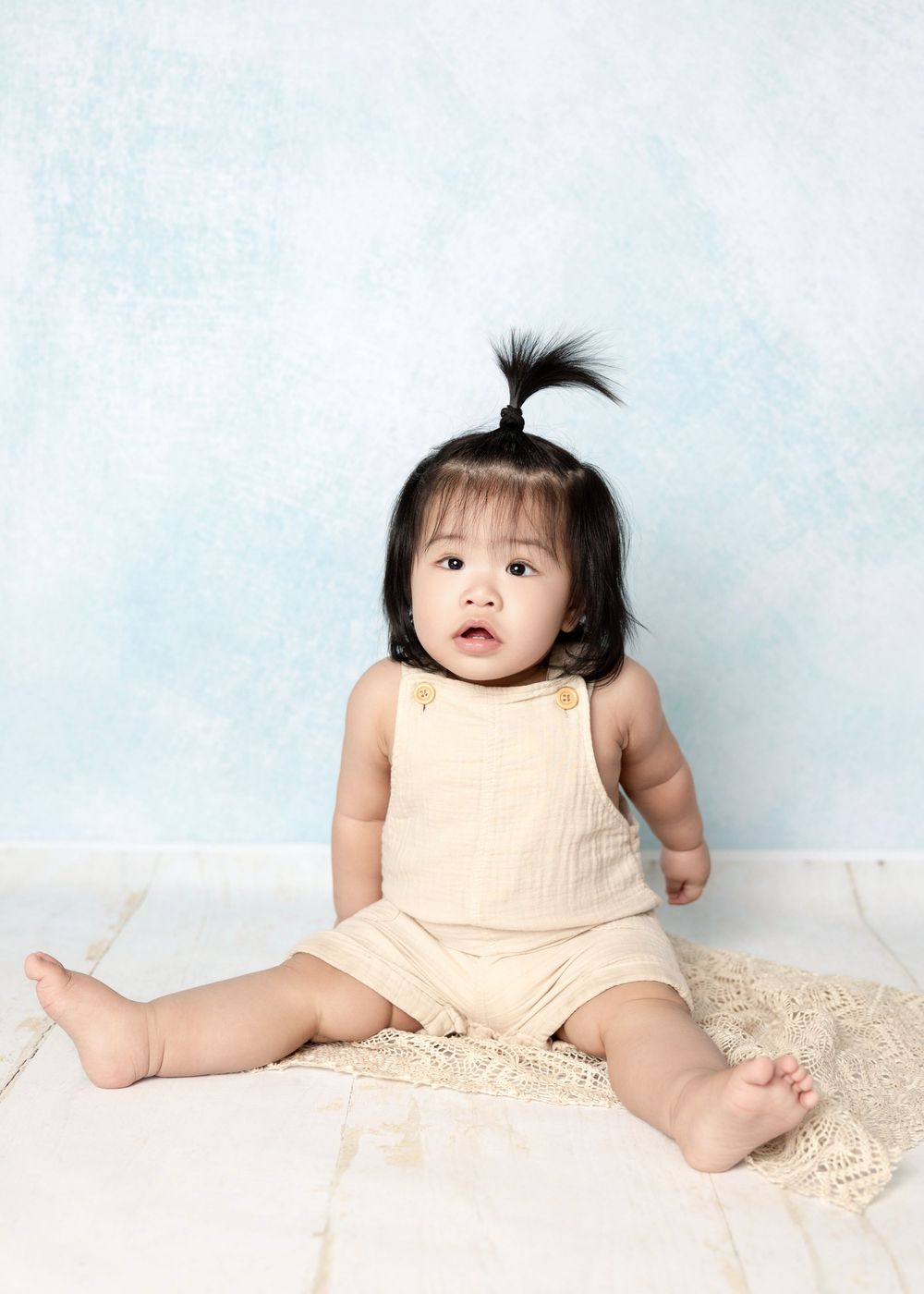 Adorable baby girl with a tiny ponytail sits in a cream romper against a soft blue backdrop.